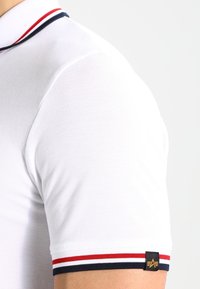 Polo shirt blanc à manches courtes avec un col et des poignets rayés rouges, blancs et bleu marine, et une petite étiquette de logo noire sur la manche.