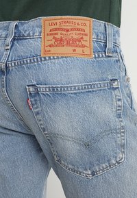 Jeans de mezclilla azul claro con una superficie texturizada, que cuentan con una etiqueta de parche de cuero marrón, dos bolsillos traseros y patrones de costura distintivos.