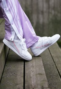 Sneaker bianchi con accenti rosa, materiale in pelle, design a bassa cima, superficie testurizzata, indossati con pantaloni sportivi lavanda dotati di bottoni a pressione.