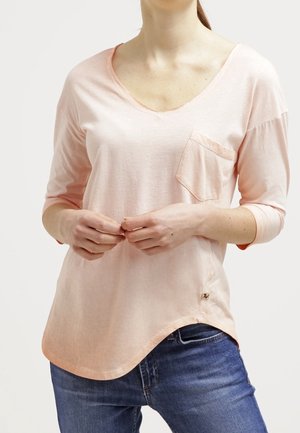 Vrouw draagt een lichtroze V-hals shirt met driekwart mouwen en een borstzak, gecombineerd met een blauwe spijkerbroek.