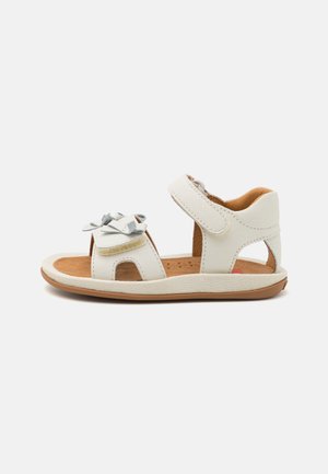 Sandalias de cuero blanco con una correa ajustable, lados con corte y un lazo decorativo. Plantilla marrón y suela de goma texturizada.
