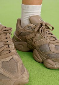 Baskets beiges avec des panneaux en daim texturé et en mesh, dotées d'une semelle épaisse, d'un bout rond et de lacets proéminents, sur un fond vert.