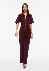 Tuta in velluto a coste color bordeaux con maniche corte, cerniera frontale, due tasche sul petto e vita con cintura, caratterizzata da pantaloni a gamba larga.