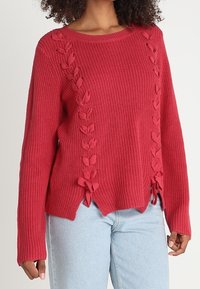 Punainen neulottu villapaita, jossa on resorikuviointi, lehtimuotoisia yksityiskohtia ja nyörit sivuilla, ja hieman väljä istuvuus.