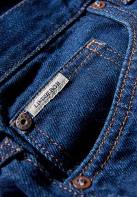 Υφασμα denim σε βαθύ μπλε χρώμα με αντιθετική ραφή σε πορτοκαλί. Διαθέτει ραμμένη ετικέτα με την ένδειξη «Lindbergh» και λεπτομέρεια με καφέ σκούρο κουμπί.