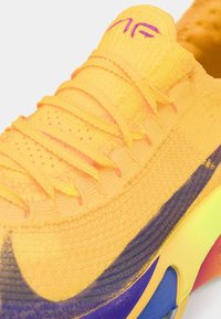 Ryškiai geltonas sportinis batelis su tekstūruotu viršumi ir purple akcentais, su plokščiomis raištelėmis, paminkštintu padu ir elegantišku dizainu.