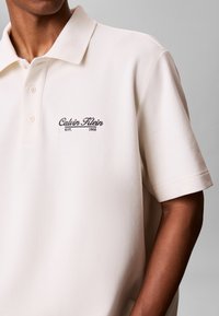 Άνδρας που φοράει εκρού πόλο μπλούζα Calvin Klein με γιακά και κουμπιά, λογότυπο με "EST. 1968" στο στήθος, εμφανές χέρι σε γκρίζο φόντο.