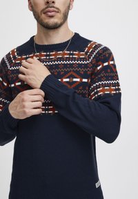 Pulover tricotat de culoare bleumarin, cu un design patterned pe piept în nuanțe de roșu, alb și maro. Are mansete cu reazem și guler rotund.