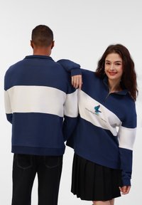 Pulover s plavim i bijelim prugama, s ovratnikom na zatvarač, s izvezenim dizajnom leptira i natpisom. Nositi s crnim hlačama i suknjom.