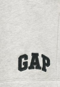 Tissu gris clair avec des lettres noires brodées "GAP" en majuscules, en gras, centrées près d'une couture surpiquée.