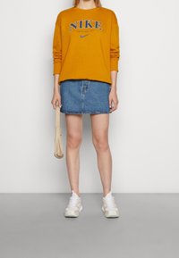 Gul sweatshirt med "NIKE"-logga, denim mini-kjol, krämfärgade sneakers och en liten beige väska. Avslappnad outfit med varierade texturer.