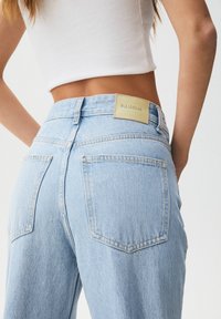 Jean en denim bleu clair avec deux poches arrière, taille mi-haute, et une étiquette de marque beige sur la ceinture. Texture lisse avec une couture classique.