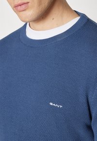 Homme portant un pull GANT bleu texturé par-dessus une chemise blanche à col rond, montrant le bas du visage et le cou sur un fond uni.