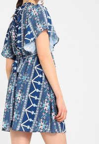 Žena v modrých květinových šatech s krátkým rukávem a zúženým pasem, stojící na pozadí v bílé barvě.