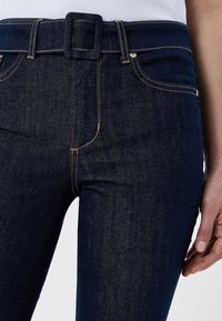 Jean denim bleu foncé avec une ceinture à boucle carrée, jambes à coupe droite, coutures contrastées et design à cinq poches.