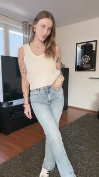 Hvit stripete tanktop, lyseblå flared jeans og svart-hvite joggesko. Rominnredning med mørkt møblement og et TV-apparat i bakgrunnen.