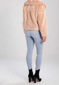 En person som bär ljusblå stretchjeans, svarta klackboots vid anklarna och en blek rosa fuskpälsjacka, står mot en neutral bakgrund.