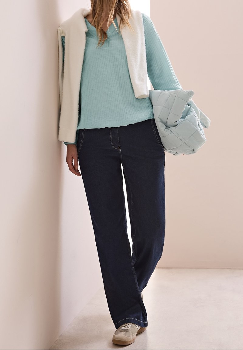 Femme portant un pull bleu clair, un pantalon bleu foncé, des chaussures beiges, avec une veste blanche posée sur les épaules et tenant une veste matelassée bleu clair.