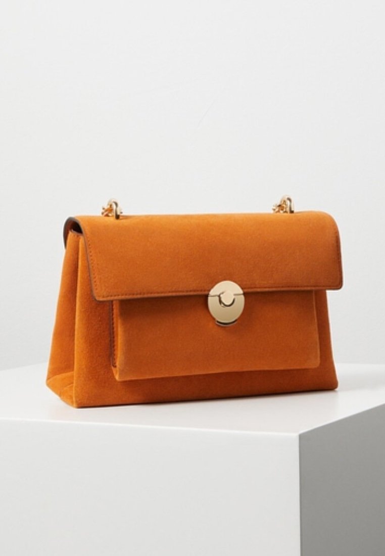 Sac à main en daim orange avec une forme structurée, un rabat supérieur et une fermeture circulaire dorée. Comprend une poche avant et une bandoulière en chaîne dorée.
