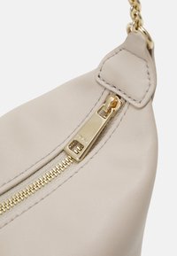 Borsa in pelle beige con superficie testurizzata, dotata di una cerniera e dettagli in oro. Il design include una tracolla a catena e un tirante a forma triangolare per la cerniera.