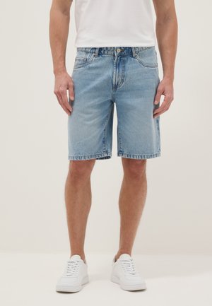 Hombre con pantalones cortos de mezclilla azul claro, zapatillas blancas y camiseta blanca, de pie contra un fondo liso y claro.