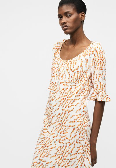 Γυναίκα που φοράει λευκό φόρεμα με πορτοκαλί λουλουδάτο σχέδιο και μανίκια μήκους μέχρι τους αγκώνες με βολάν στα μανίκια, στέκεται μπροστά σε απλό φωτεινό φόντο.