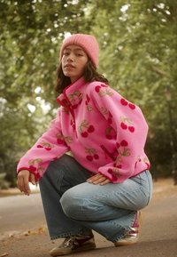 Rosa Fleece-Pullover mit Kirschmotiven, rotem Reißverschluss und gerippten Bündchen; kombiniert mit hellblauen weit geschnittenen Jeans und goldenen Sneakers.