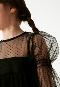 Schwarzes Kleid mit durchsichtigen, gepunkteten Tüllärmeln, festem Oberteil, gerafftem Rock und elastischen Bündchen mit weicher Textur.