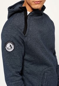 Ciemnoniebieska bluza z kapturem wykonana z teksturowanej tkaniny, z bocznym zamkiem, przednimi kieszeniami oraz okrągłą naszywką z logo na lewym ramieniu.