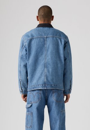 Homme portant une veste en denim bleu clair à fines rayures et un jean assorti, debout de dos sur un fond uni.