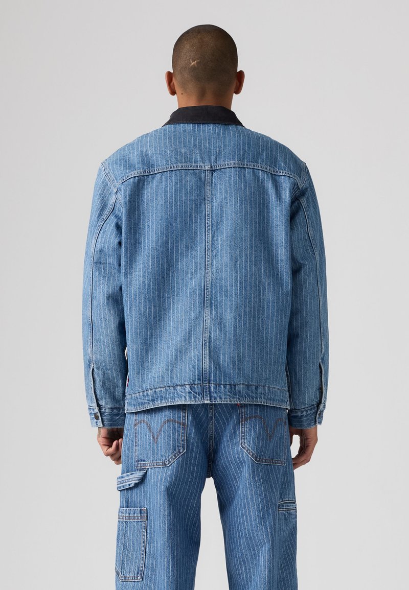 Homme portant une veste en denim bleu clair à fines rayures et un jean assorti, debout de dos sur un fond uni.