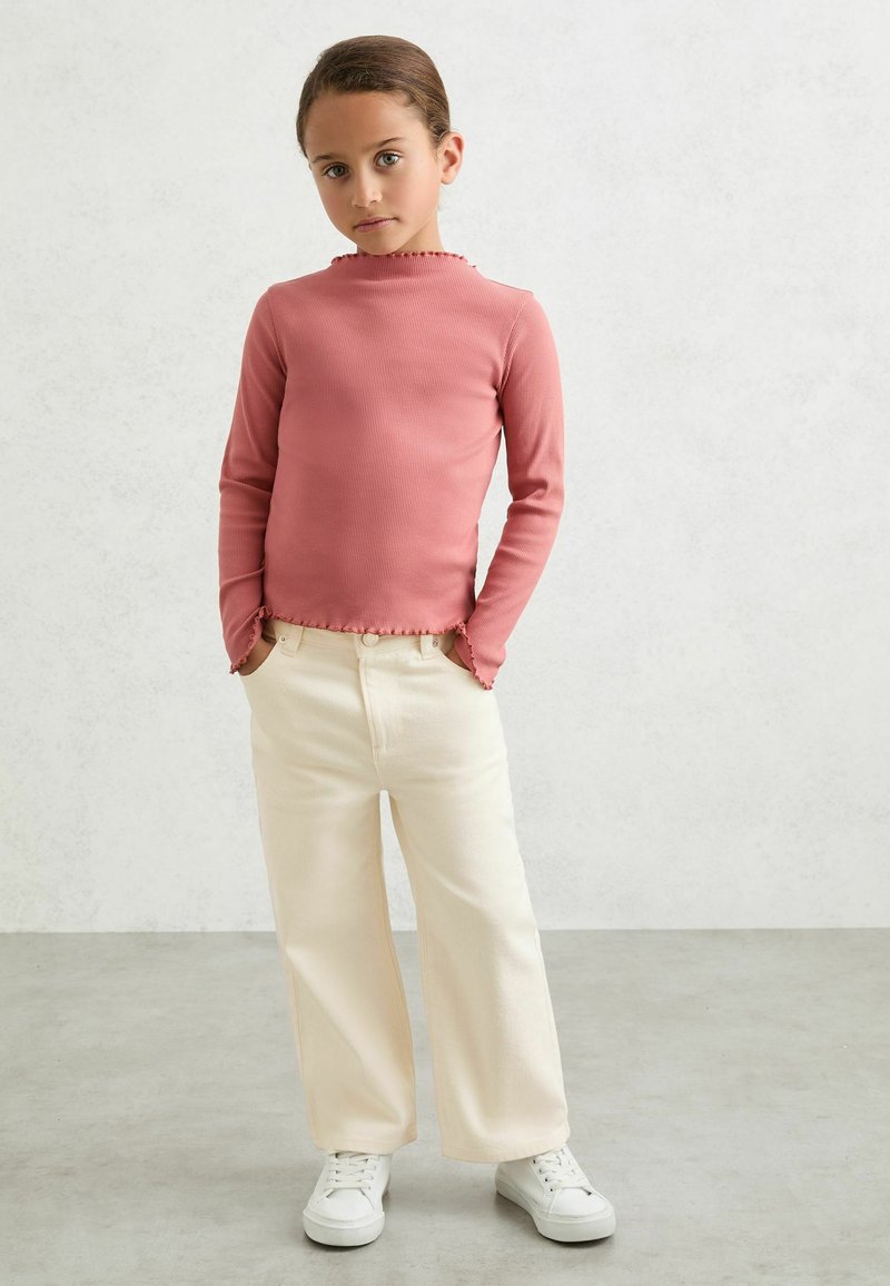 Haut côtelé à manches longues couleur rose poussière avec un col à volants, associé à un pantalon large crème et des baskets blanches. Design simple, coupe décontractée.