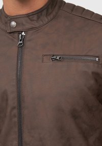 Veste en cuir synthétique marron avec un col montant, un zip à l'avant et une poche poitrine zippée. Présente des détails aux épaules et une texture lisse.