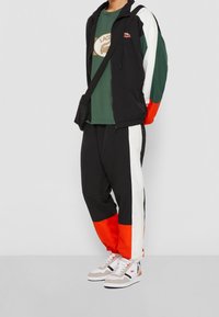 Träningsjacka i svart, grön och vit; matchande byxor med orange detaljer. Vita sneakers med grå mocka och svarta ränder. Avslappnad atletisk stil.