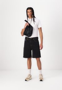 Sorte denimshorts med gule stikninger, parret med en hvid T-shirt og beige sneakers. En sort skuldertaske med lynlås bæres i den ene hånd.