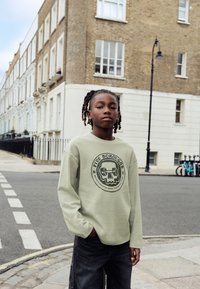 Šviesiai žalias ilgomis rankovėmis megztinis su apvaliu grafiniu spaudiniu priekyje, derinamas su tamsiais džinsais. Atsitiktinė miesto gatvių aplinka.