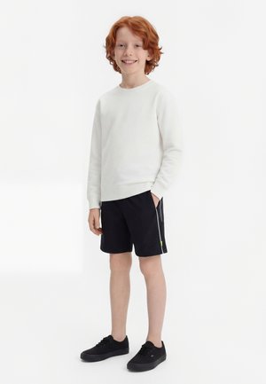 Rødhåret dreng smilende, iført hvid langærmet skjorte, sorte shorts med hvid stribe og sorte sneakers, stående med den ene hånd i lommen.