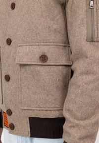 Beige Wolljacke mit Knopfklappen-Tasche, braunen Knöpfen, schwarzem geripptem Bündchen, Reißverschlusstasche am Ärmel und orangefarbigem Aufnäher nahe dem Saum.