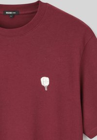 T-shirt en coton bourgogne avec un col rond et des manches courtes. Présente un petit graphique blanc ressemblant à une pagaie avec du texte.