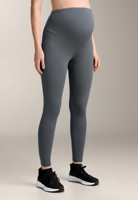 Nosečnostne legice v temno sivi barvi, visok pas, gladka tekstura, oprijet kroj, v kombinaciji s črnimi športnimi čevlji.