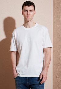 Hvid bomulds T-shirt med rund halsudskæring, korte ærmer, der har et lille prægede logo på brystet. Matchet med blå denimjeans.