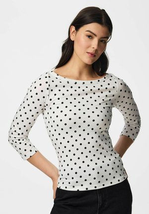 Jeune femme aux cheveux foncés portant une chemise blanche à manches longues à pois noirs, debout les mains sur les hanches devant un fond uni.