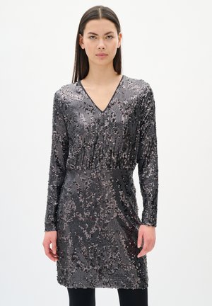 Eine Frau trägt ein enganliegendes, langärmliges Kleid aus dunkelsilbernen Pailletten mit V-Ausschnitt und steht vor einem schlichten weißen Hintergrund.