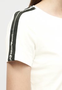 Λευκό κοντομάνικο πουκάμισο με μαύρη υφασμάτινη λωρίδα στον ώμο που φέρει την επιγραφή «EA7 Emporio Armani» σε λευκό κείμενο.