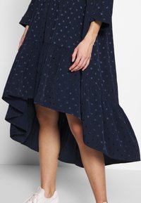 Mörkblå klänning med prickigt mönster; har en lager-på-lager, asymmetrisk nederkant och flödande tyg. Bärs med vita sneakers.