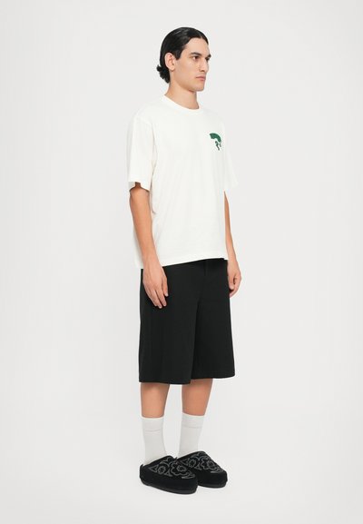 T-shirt de algodão branca com gráfico verde, combinada com calções pretos até ao joelho e sapatos slip-on pretos bordados, complementados por meias brancas.