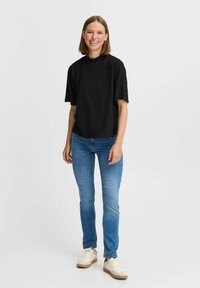 Sort ribbet t-shirt med korte ærmer, løstsiddende pasform, parret med lyseblå slim-fit jeans og beige sko med hvide detaljer. Ensfarvet baggrund.