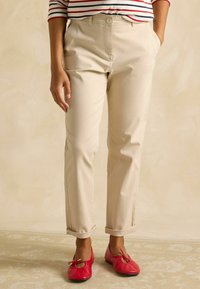 Bēša toņa čino bikses ar piegulošu piegriezumu, sānu kabatām un uzlocītiem galiņiem, kombinētas ar sarkaniem dzīvokļiem. Smalka tekstūra, neitrāla krāsa.