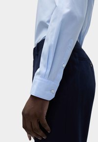 Camicia a maniche lunghe azzurro chiaro con polsini a bottone, abbinata a pantaloni blu scuro. Il tessuto è liscio con una leggera lucentezza. I polsini presentano un singolo bottone.