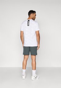 Λευκή μπλούζα με καμουφλάζ μοτίβο, με μαύρη ετικέτα "BORG" στο πίσω μέρος, συνδυασμένη με σκούρο πράσινο σορτς και λευκά αθλητικά παπούτσια με κάλτσες.